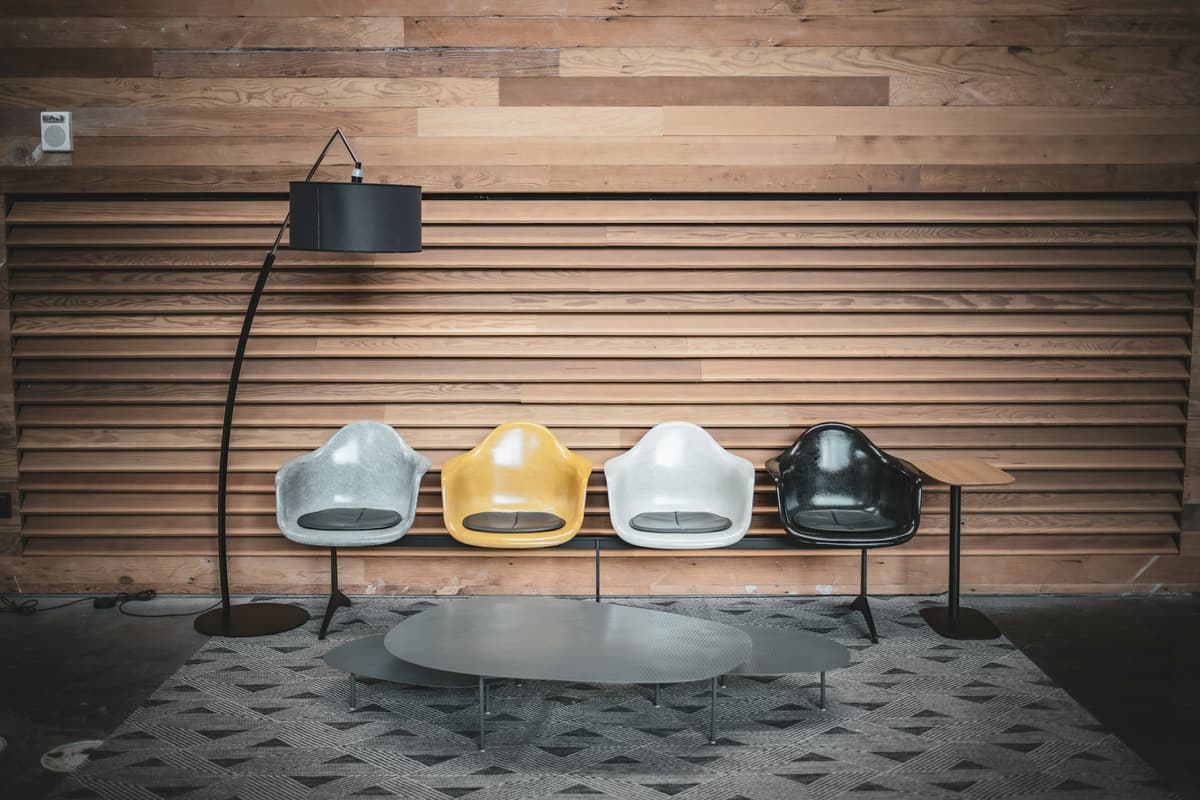 Wartebereich mit vier Schalenstühlen vor einer Holzwand, Stehlampe und kleinem Tisch